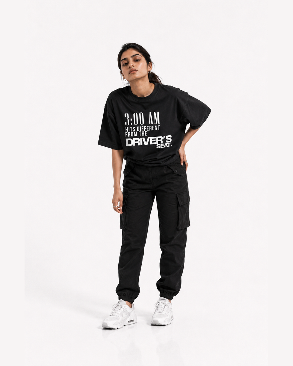 A woman modeling a black oversized graphic T-shirt with white text reading '3:00 AM HITS DIFFERENT FROM THE DRIVER'S SEAT.' paired with black cargo pants and white sneakers, against a plain light gray studio background.