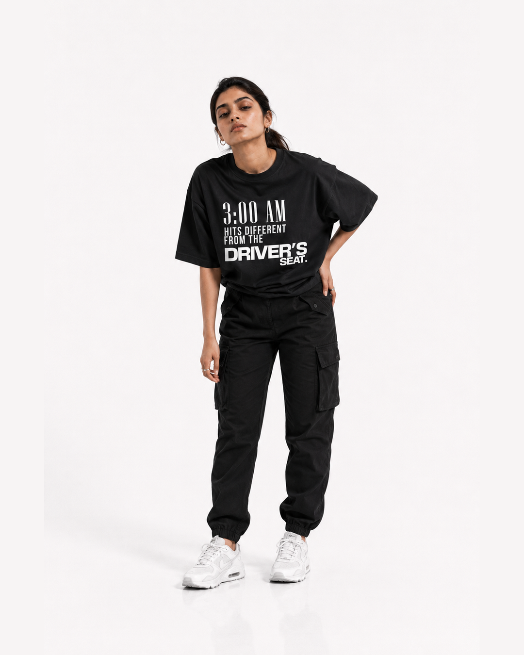 A woman modeling a black oversized graphic T-shirt with white text reading '3:00 AM HITS DIFFERENT FROM THE DRIVER'S SEAT.' paired with black cargo pants and white sneakers, against a plain light gray studio background.