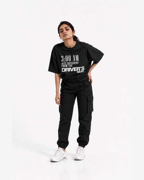 A woman modeling a black oversized graphic T-shirt with white text reading '3:00 AM HITS DIFFERENT FROM THE DRIVER'S SEAT.' paired with black cargo pants and white sneakers, against a plain light gray studio background.