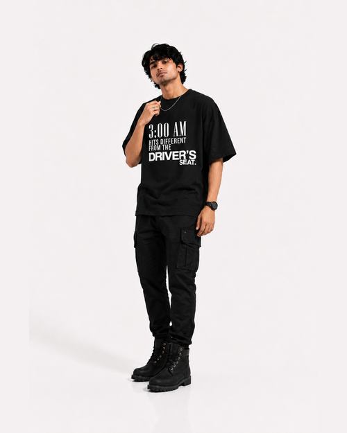 A man modeling a black graphic t-shirt with white text that reads '3:00 AM HITS DIFFERENT FROM THE DRIVER'S SEAT', paired with black cargo pants and black boots, against a plain light gray background.