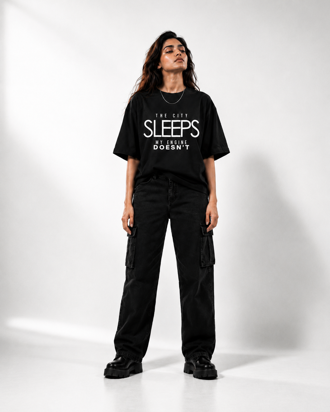 A woman modeling a black graphic t-shirt with white text that reads 'THE CITY SLEEPS MY ENGINE DOESN'T', paired with black cargo pants and black shoes, set against a plain white studio background.