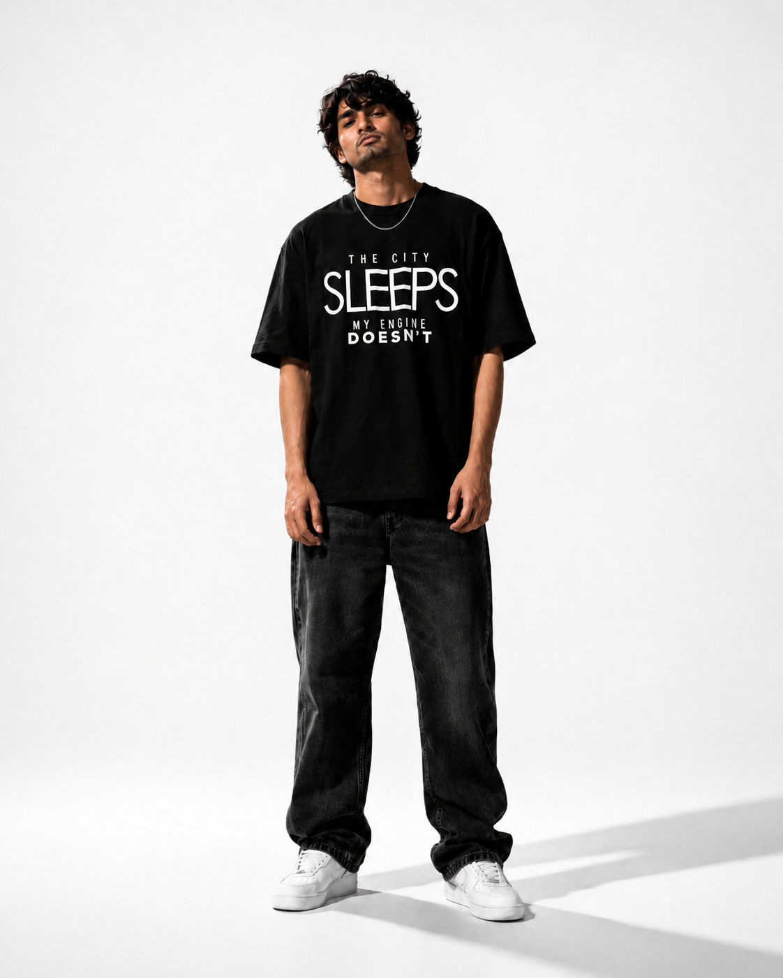 A man modeling a black graphic t-shirt with the text 'THE CITY SLEEPS BUT My ENGINE DOESN'T' in white, paired with dark gray jeans and white sneakers, shot against a plain white studio background.