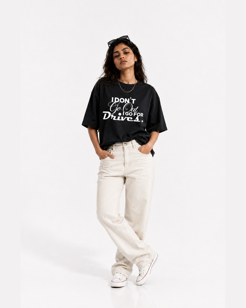 A woman modeling a black graphic t-shirt with white text that reads 'I DON'T GO OUT, I GO FOR Drives.' paired with off-white jeans and white sneakers, set against a plain white studio background.