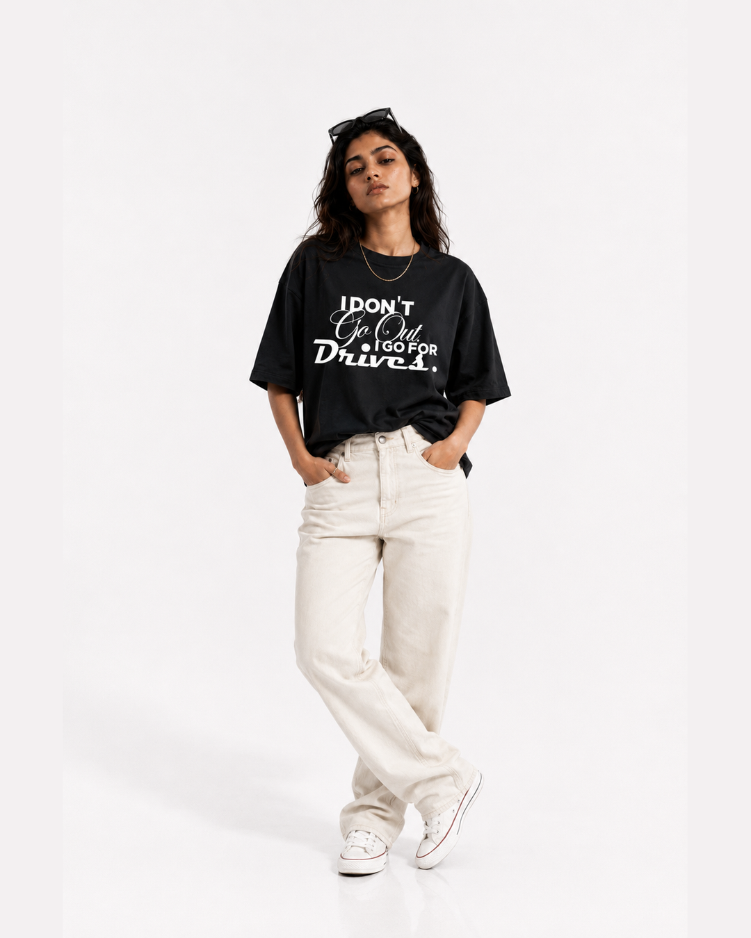 A woman modeling a black graphic t-shirt with white text that reads 'I DON'T GO OUT, I GO FOR Drives.' paired with off-white jeans and white sneakers, set against a plain white studio background.