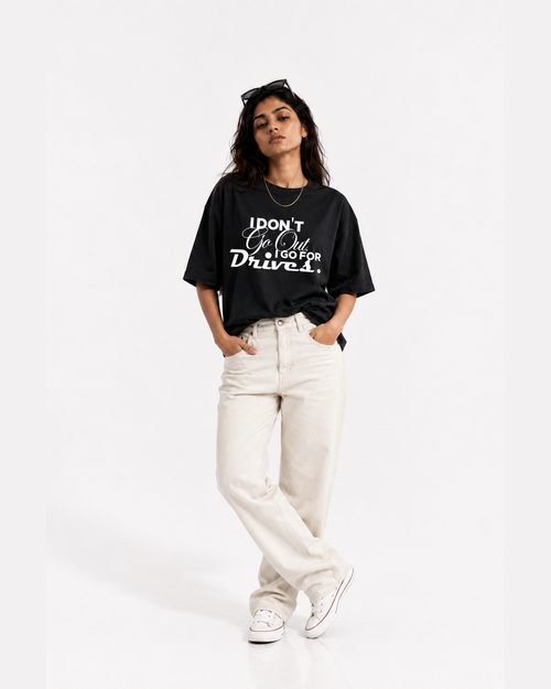 A woman modeling a black graphic t-shirt with white text that reads 'I DON'T GO OUT, I GO FOR Drives.' paired with off-white jeans and white sneakers, set against a plain white studio background.