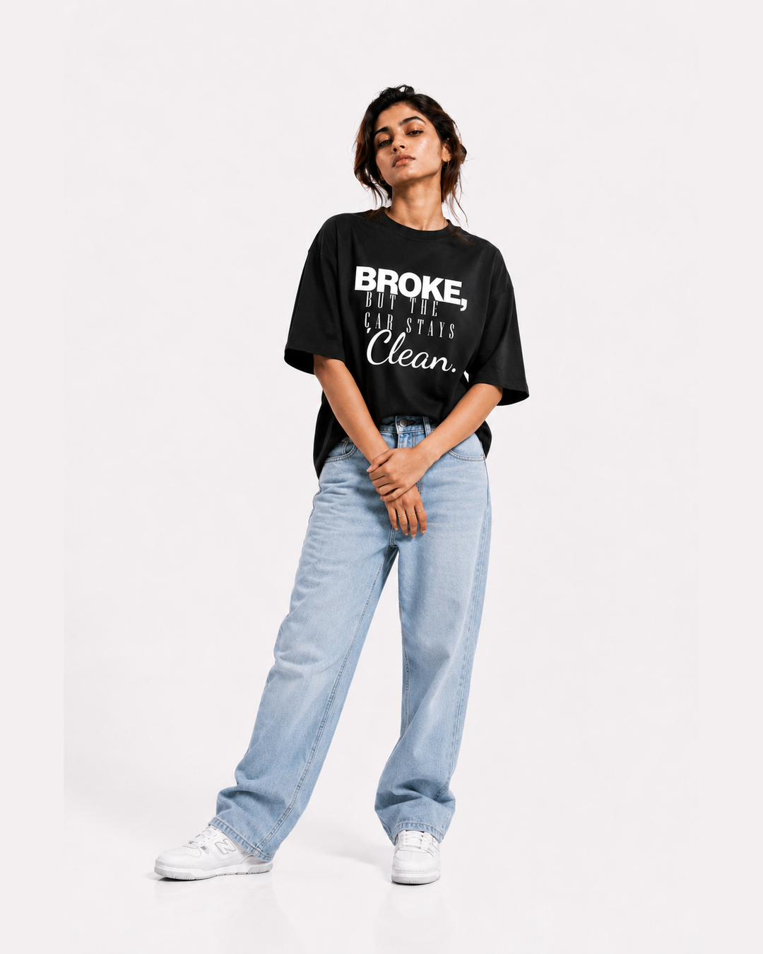 A woman modeling a black graphic t-shirt with white text that reads 'BROKE, BUT THE CAR STAYS CLEAN.