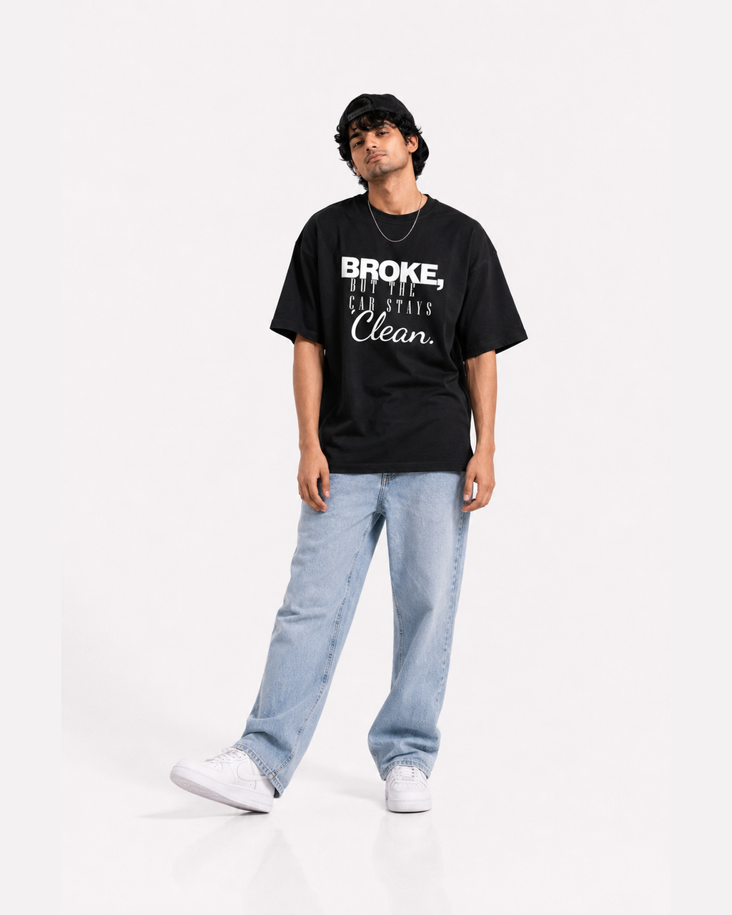 A man modeling a black graphic t-shirt with white text that reads 'BROKE, BUT THE CAR STAYS Clean.' paired with light blue jeans and white sneakers on a white studio background.