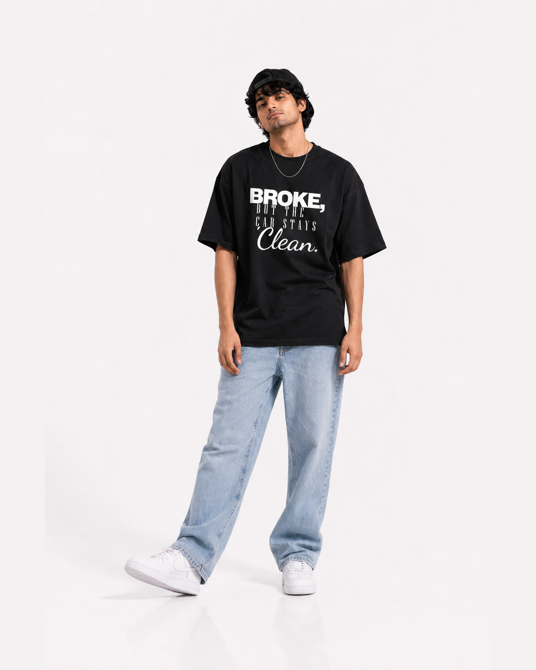 A man modeling a black graphic t-shirt with white text that reads 'BROKE, BUT THE CAR STAYS Clean.' paired with light blue jeans and white sneakers on a white studio background.
