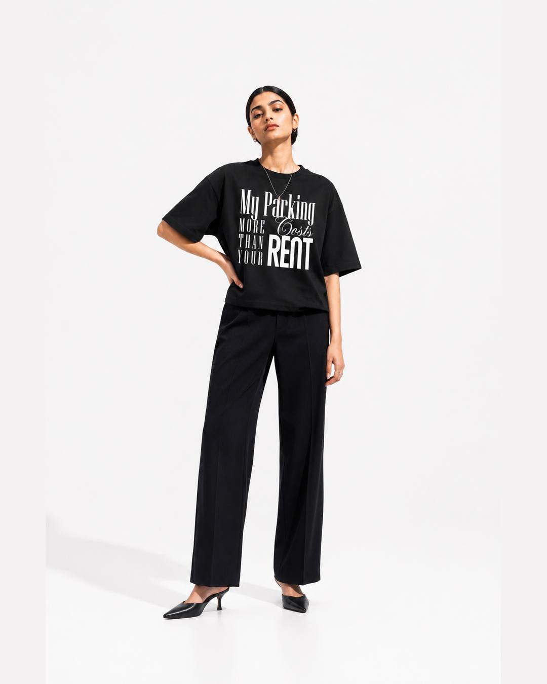 A woman modeling a black graphic t-shirt and black wide-leg trousers against a clean white studio background. The t-shirt features white text that reads 'My Parking COSTS MORE THAN YOUR RENT'.