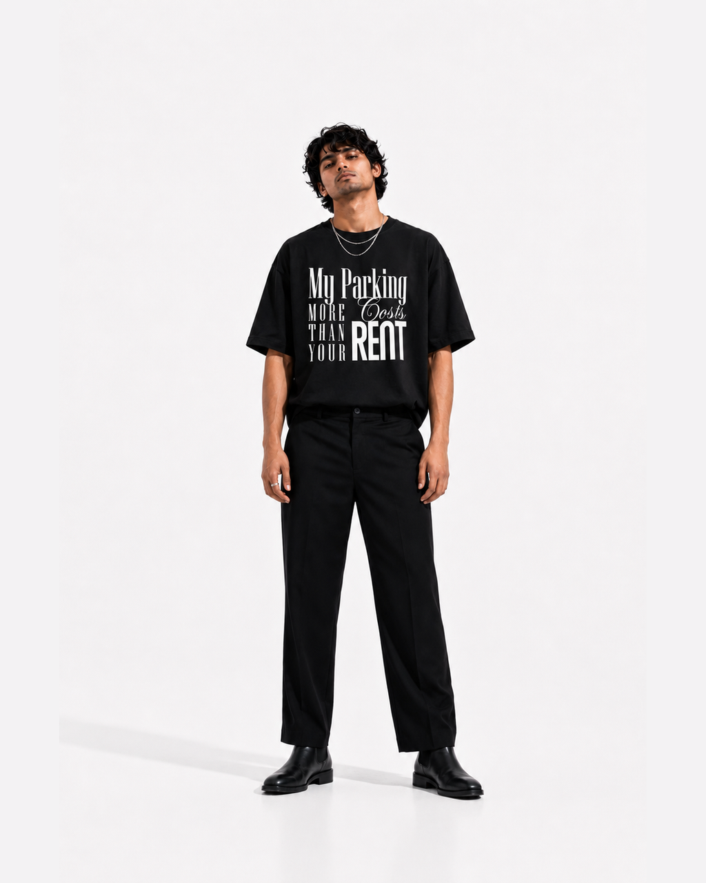 A man modeling a black graphic t-shirt with white text that reads 'My Parking COSTS MORE THAN YOUR RENT', paired with black trousers and black shoes, against a plain white studio background.