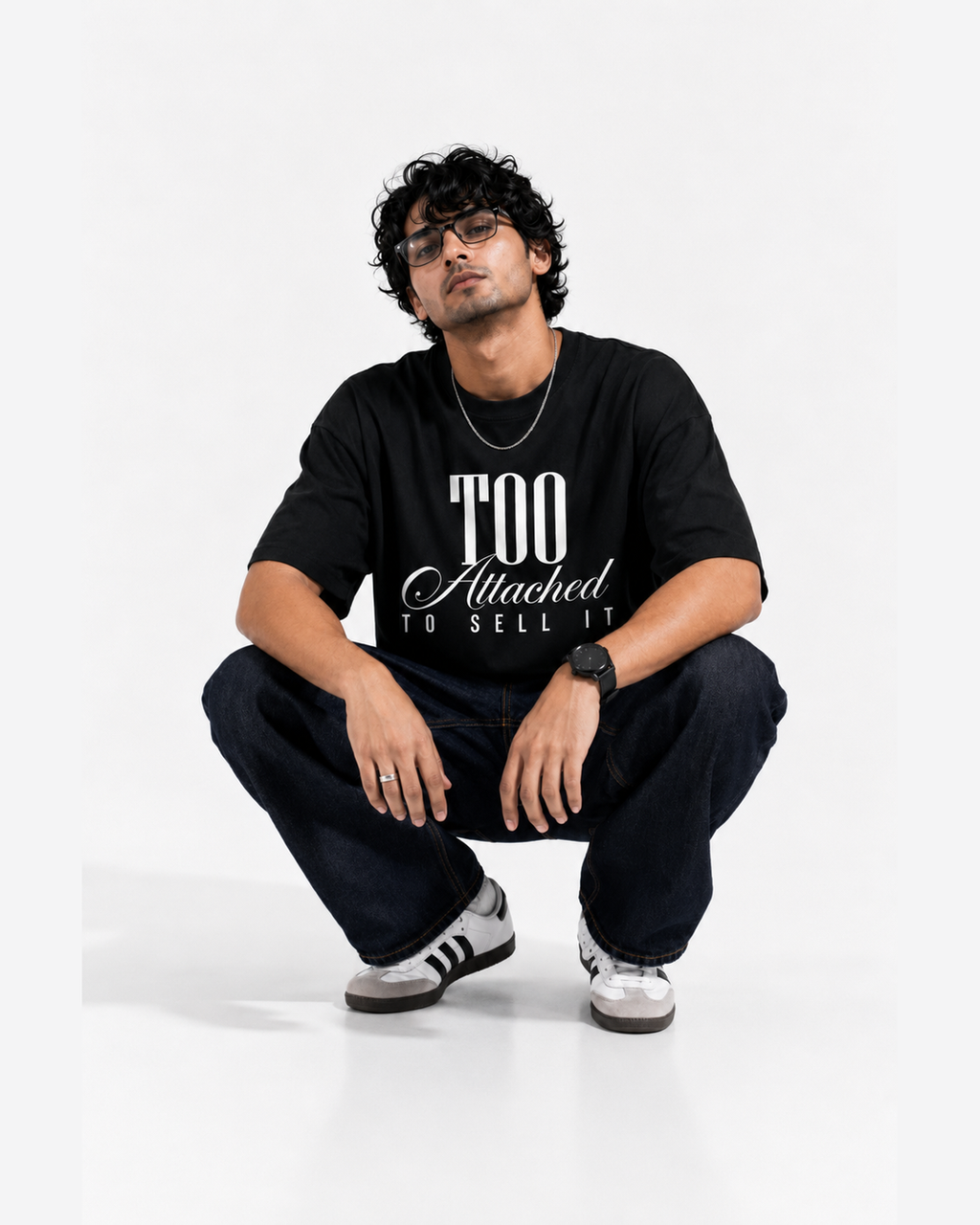 A man wearing a black graphic t-shirt with the text 'TOO Attached TO SELL IT' in white, paired with dark jeans and white sneakers, crouching against a plain white studio background.