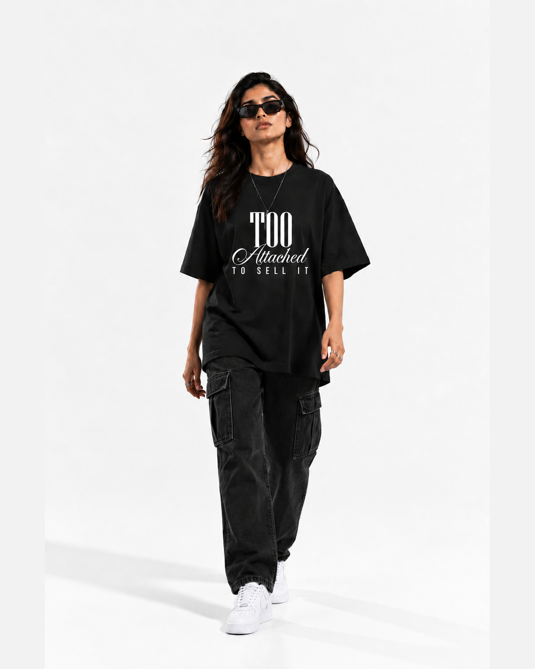 A woman modeling an oversized black graphic T-shirt with white text reading 'Too Attached To Sell It', paired with dark cargo pants and white sneakers, against a plain white studio background.