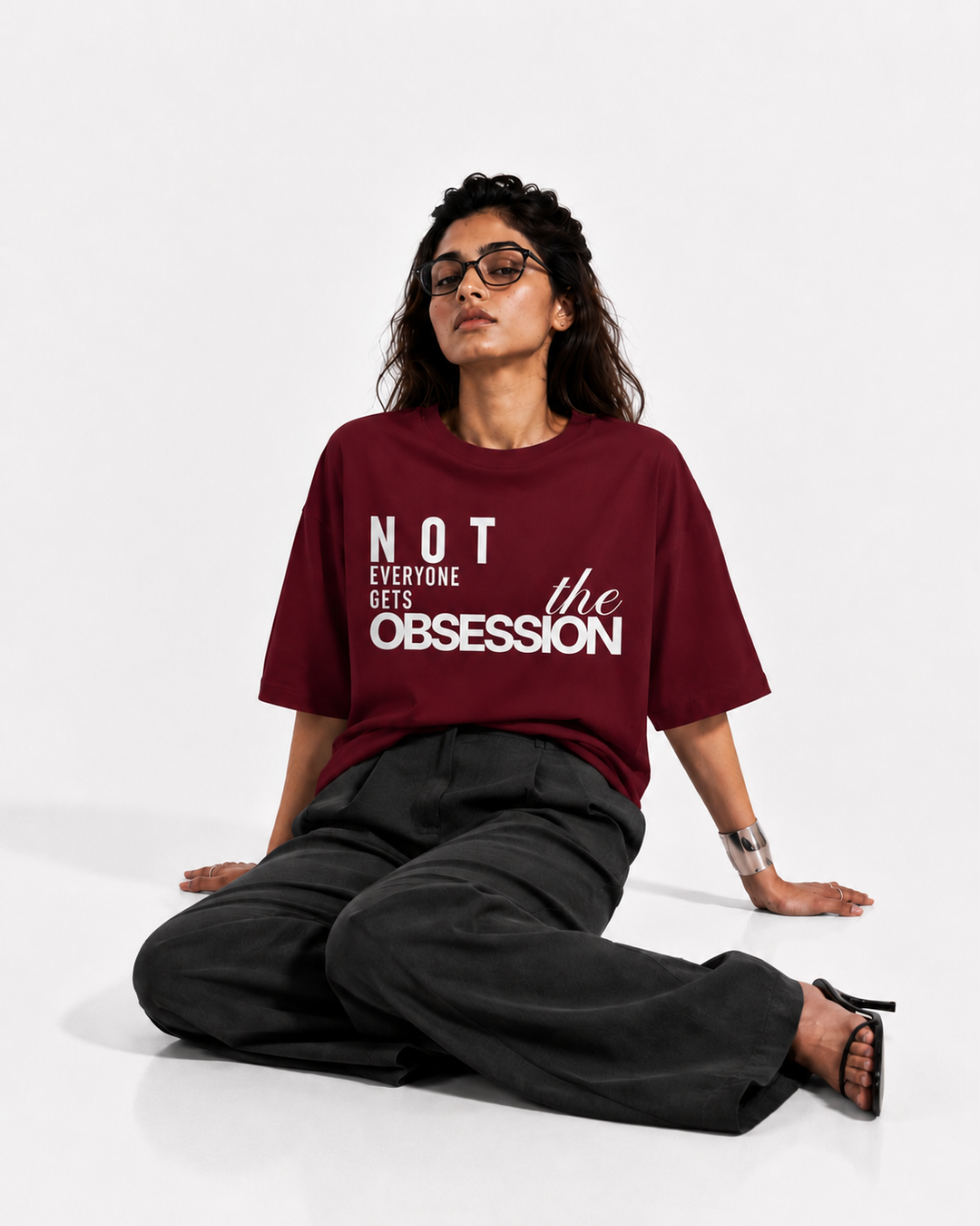 A woman wearing a maroon short-sleeve graphic t-shirt with the text 'NOT EVERYONE GETS THE OBSESSION' in white, paired with black wide-leg trousers and black sandals, sitting on a white studio floor.