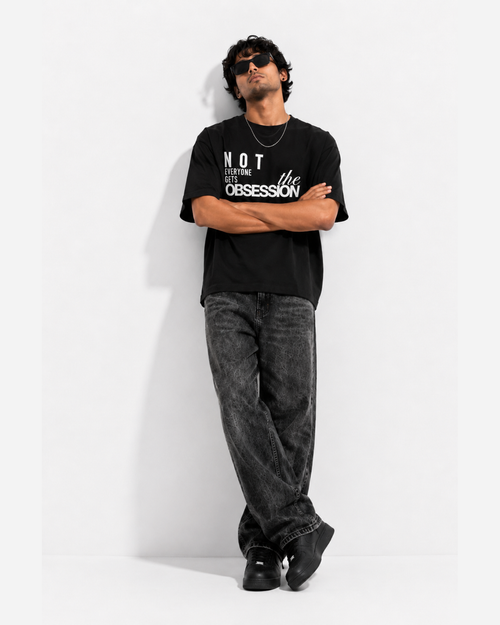 A man wearing a black graphic t-shirt with the text 'NOT everyone gets the OBSESSION' in white, paired with dark gray washed jeans and black shoes, leaning against a white wall.