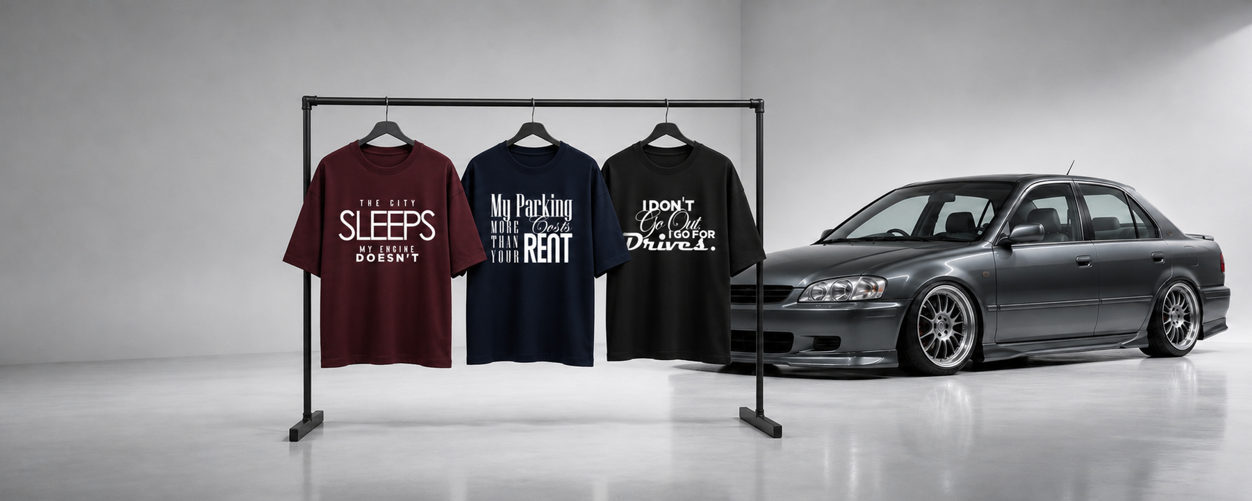 Three graphic t-shirts in maroon, navy, and black hang on a clothing rack next to a dark gray customized sedan in a minimalist studio setting.