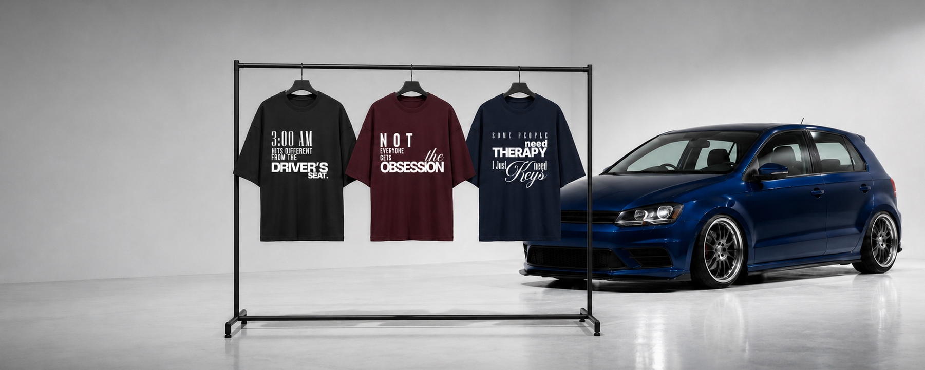 Three graphic T-shirts displayed on a black clothing rack next to a dark blue Volkswagen Golf in a studio setting.